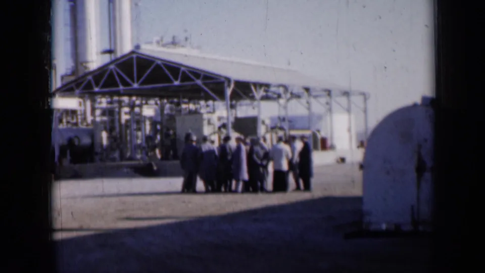 1961 Missouri 8mm frame showing men and women gathered beneath a metal canopy beside an industrial building and storage tank.