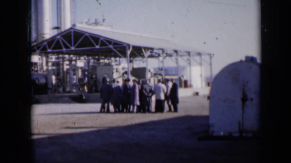 1961 Missouri 8mm frame showing adults gathered in the yard of an industrial site, with a canopy, silos, and rounded tank visible.