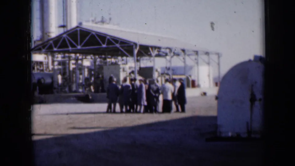 Adults in mid-century clothing stand near an industrial complex in metadata-supported 1961 Missouri footage.