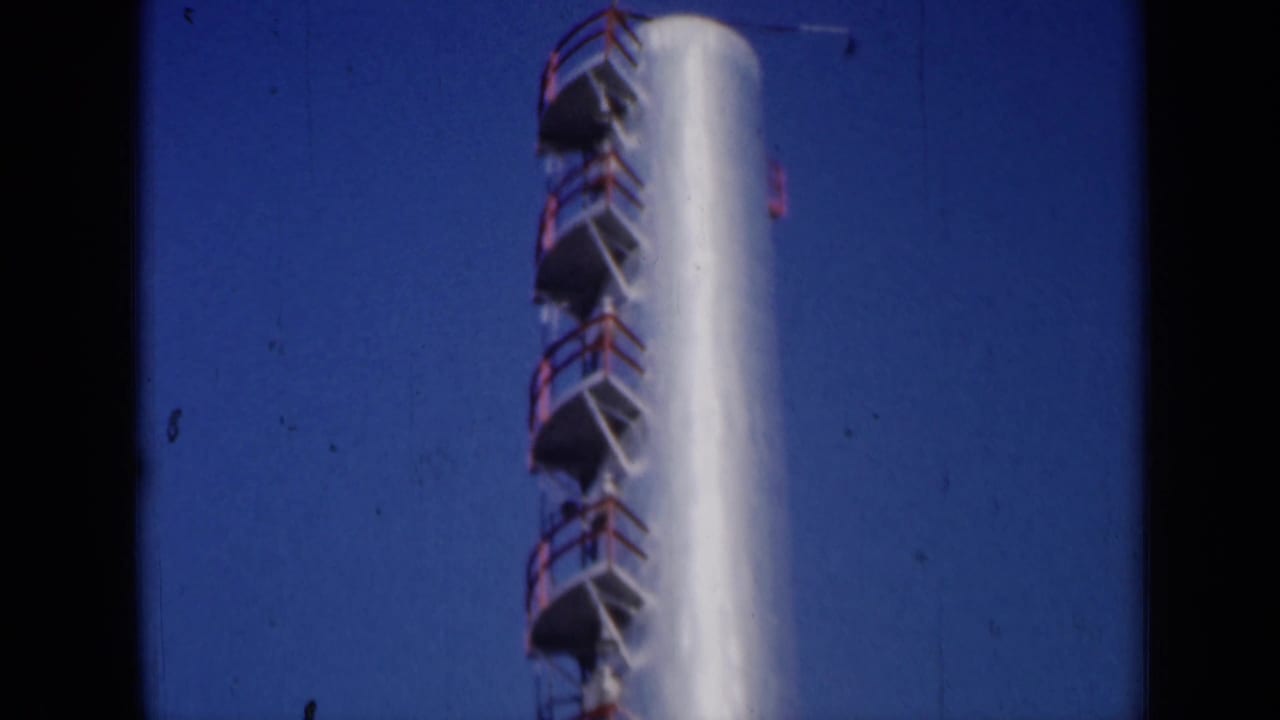 1961 Missouri 8mm view of a white industrial stack with red ladder platforms rising against a deep blue sky.