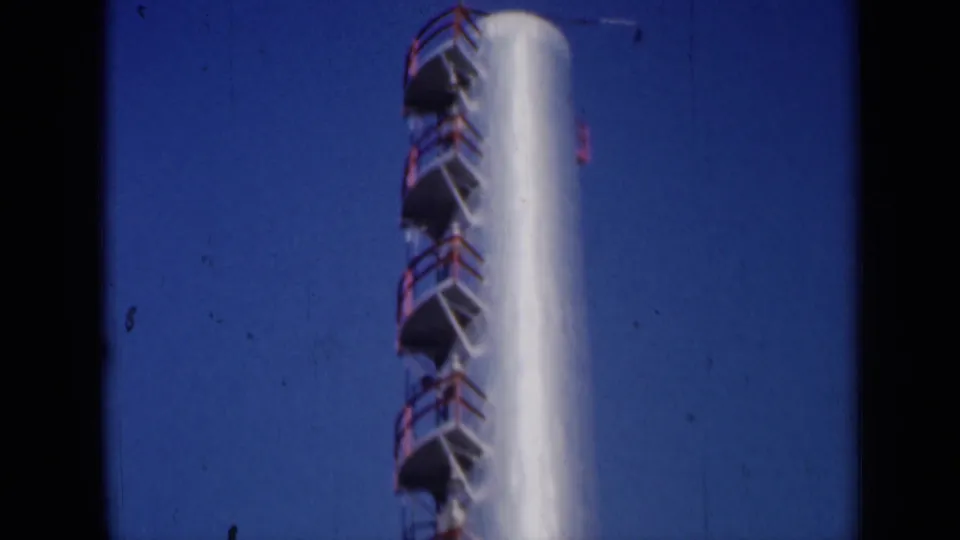 1961 Missouri 8mm view of a white industrial stack with red ladder platforms rising against a deep blue sky.