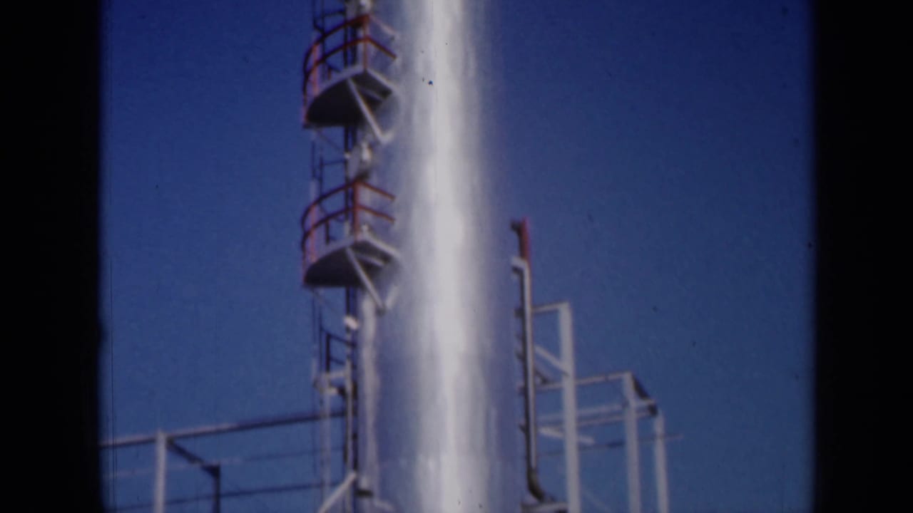 Upper section of refinery tower with platforms, railings, and pipe framework.