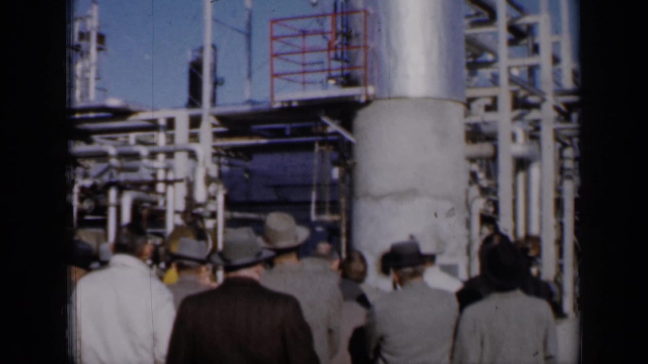 Suited visitors seen from behind while touring pipes and platforms at an industrial facility.