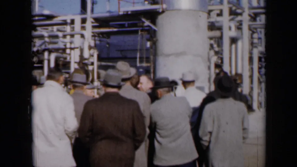1961 Missouri home-movie frame of suited visitors pausing near machinery and platforms at an industrial plant.