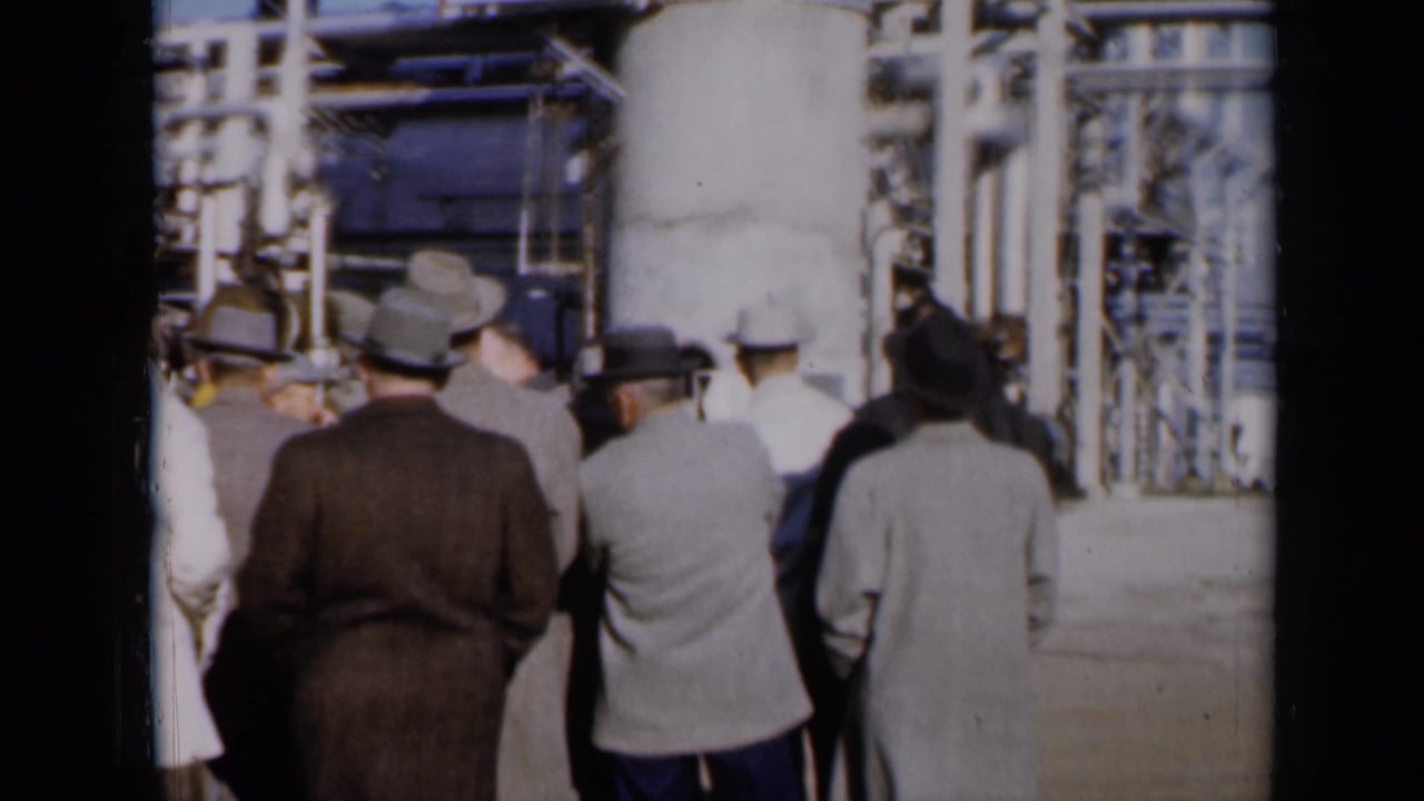 Men in hats walk near pipes and plant machinery in a sunlit industrial area.