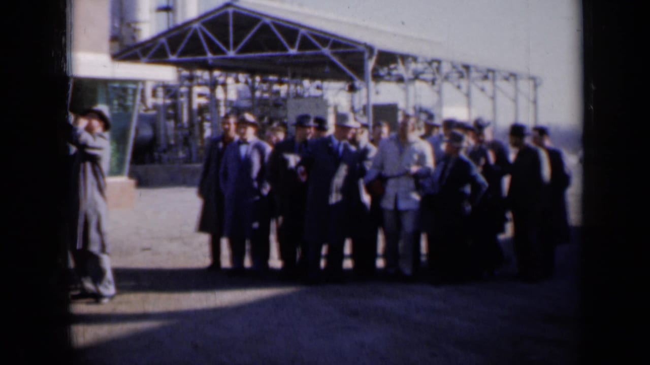 A group of men stands under a metal canopy at an industrial facility.