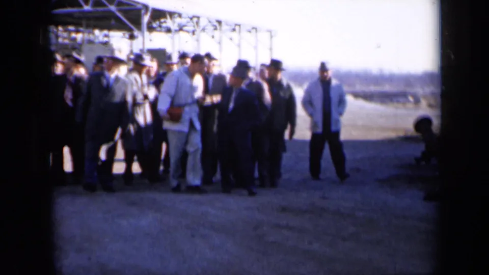 A group of men remains near the plant structures while one man walks across the sunlit pavement in the 1961 Missouri industrial scene.