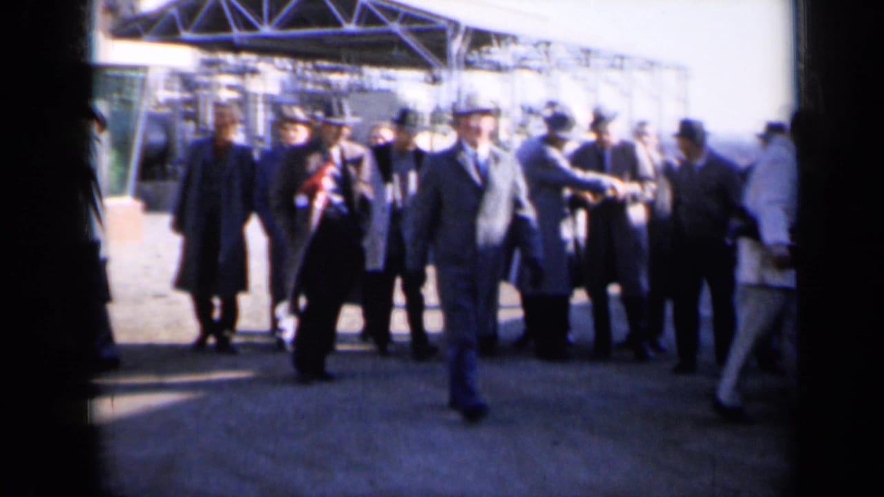 Men in suits walking and standing outside beside industrial metalwork.