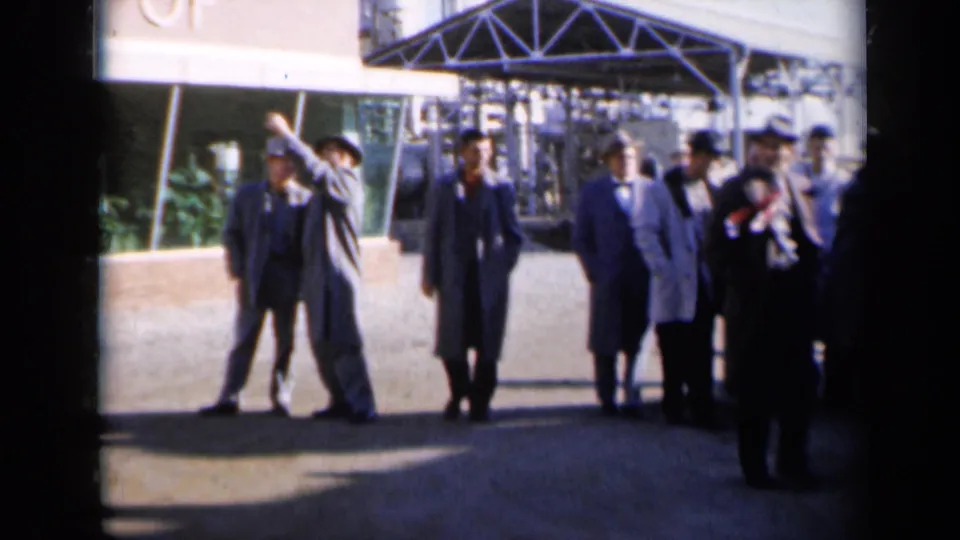 At a Missouri industrial site, one suited man gestures while others in hats and coats stand nearby in 1961 home-movie footage.
