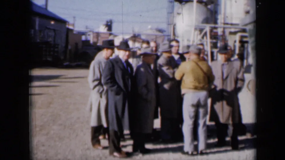 A cluster of men in business clothing gathers in a Missouri industrial yard, with buildings and equipment visible behind them.