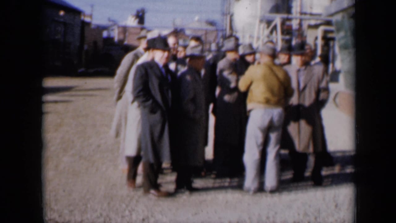 Men in coats and hats stand in a group outside an industrial plant.