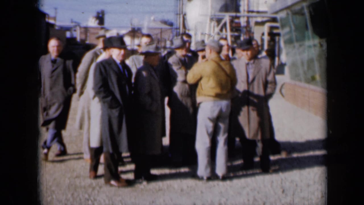 Several men stand near pipes and plant buildings while talking outdoors.
