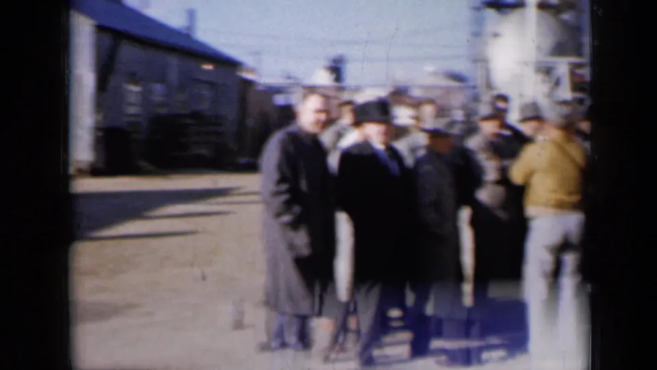 The handheld 8mm frame shows men gathered outside a Missouri plant, with pipes and buildings behind them.