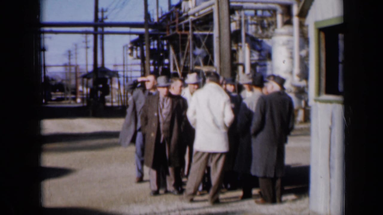 Men in overcoats and hats stand near industrial structures, seen through a dark doorway edge.