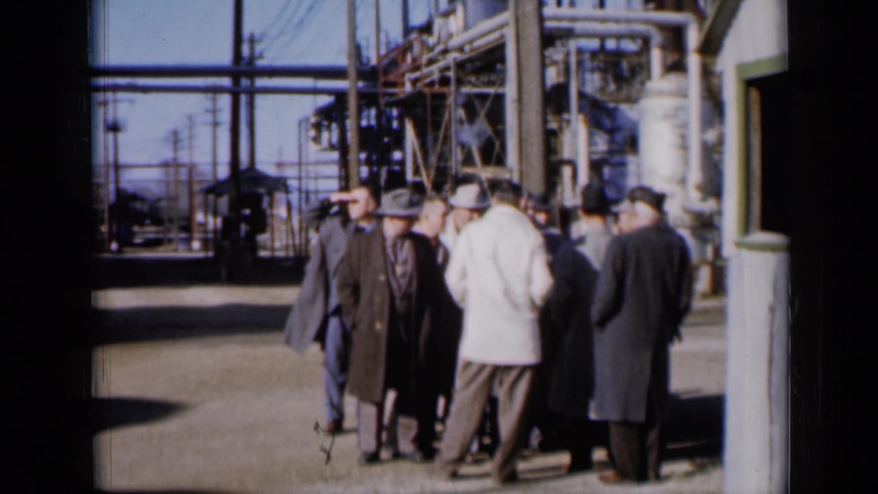 A group of men in coats and hats stands in a plant yard with pipes and structures behind them.