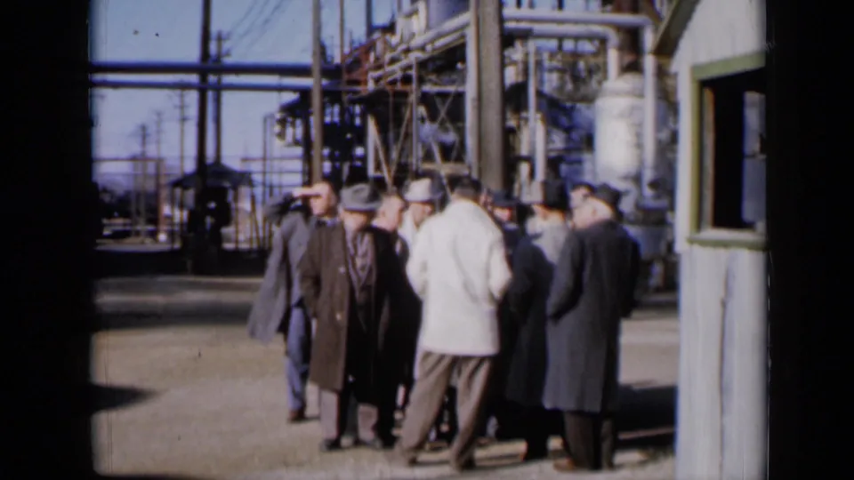 The 1961 Missouri 8mm shot holds on men in coats and fedoras gathered outside an industrial facility under clear daylight.