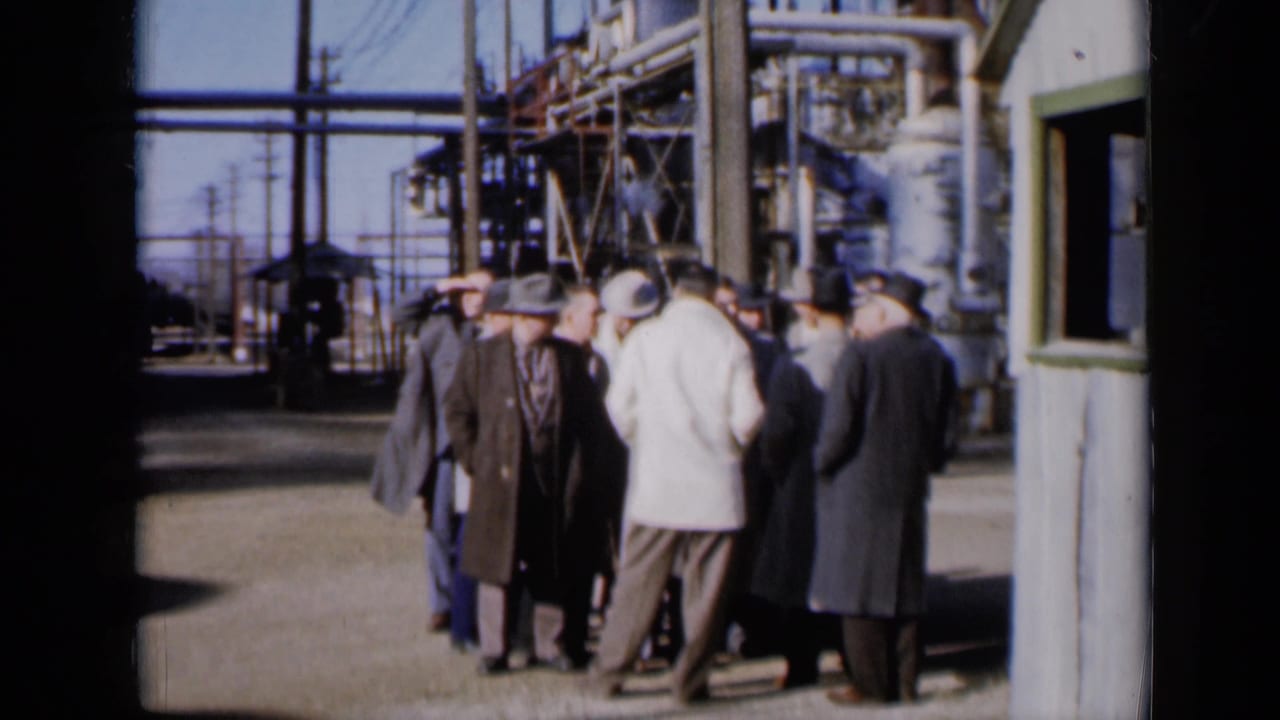 Men in suits and hats gather beside a small grey shack at an industrial plant.