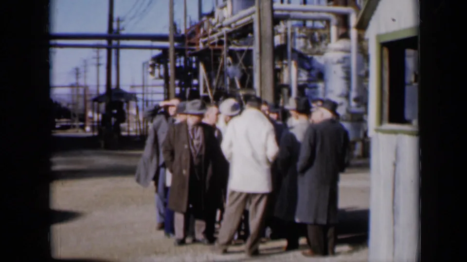 In 1961 Missouri footage, men in suits and fedoras stand outside near a grey shack, pipes, and utility structures at a factory site.