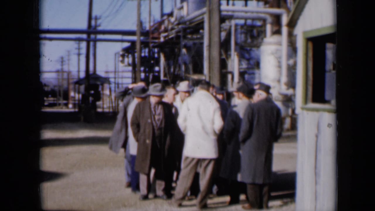 The 1961 Missouri 8mm frame shows men in dark and light overcoats clustered near factory pipework and a small grey structure.