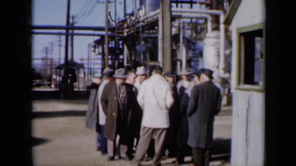 Men in 1960s suits and hats stand outdoors at a Missouri industrial plant, with a grey shack and dense pipe structures behind them.