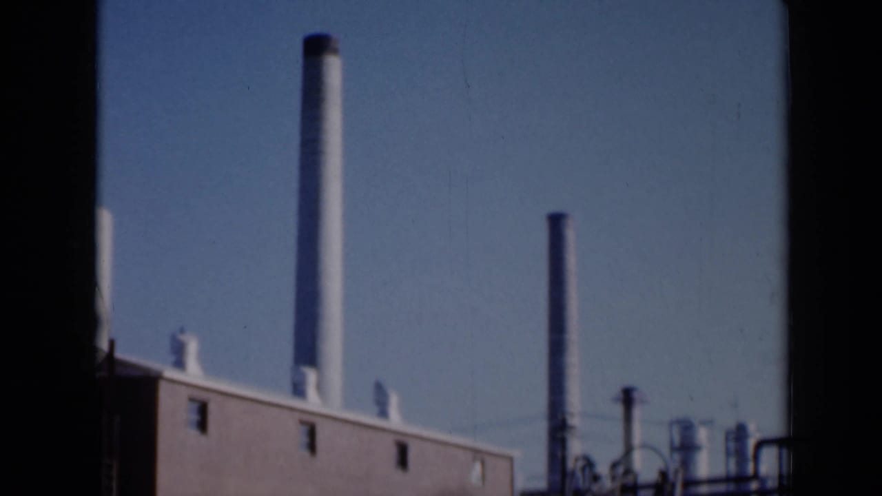 1961 Missouri 8mm view of a brick industrial roofline, white chimney stack, darker stacks and pipework under blue sky.