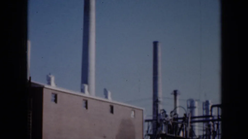 1961 Missouri 8mm frame with a brick industrial wall, pale smokestack, darker stack and rooftop vents seen through dark side framing.