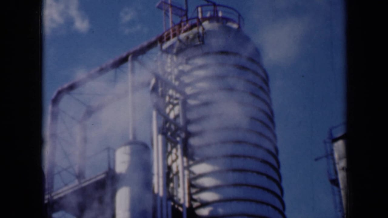 Ribbed cylindrical industrial tower with pipes, platform rails, and steam against a blue sky.
