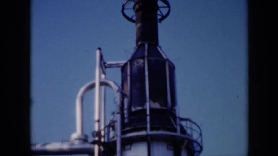 1961 Missouri home-movie frame showing a tall cylindrical industrial tower with railings, pipes and a dark upper housing against clear sky.