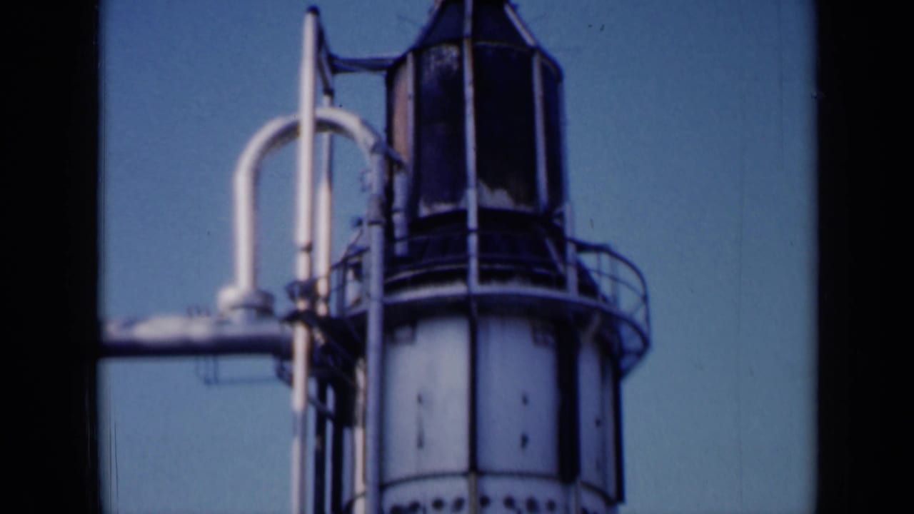 Upper portion of a cylindrical industrial tower with railings, pipes and blue sky.