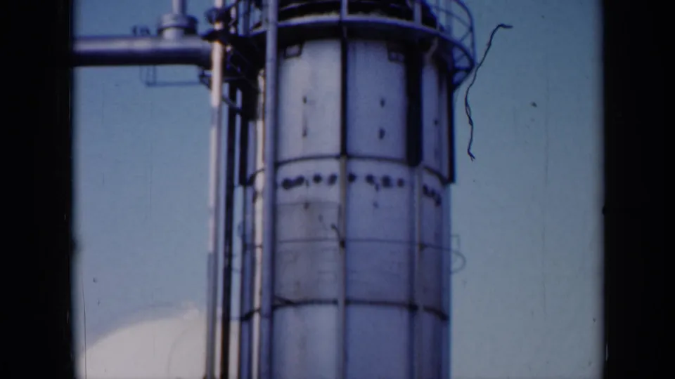 1961 Missouri 8mm frame showing a tall white industrial tank, exterior pipework, and railings at a factory site.