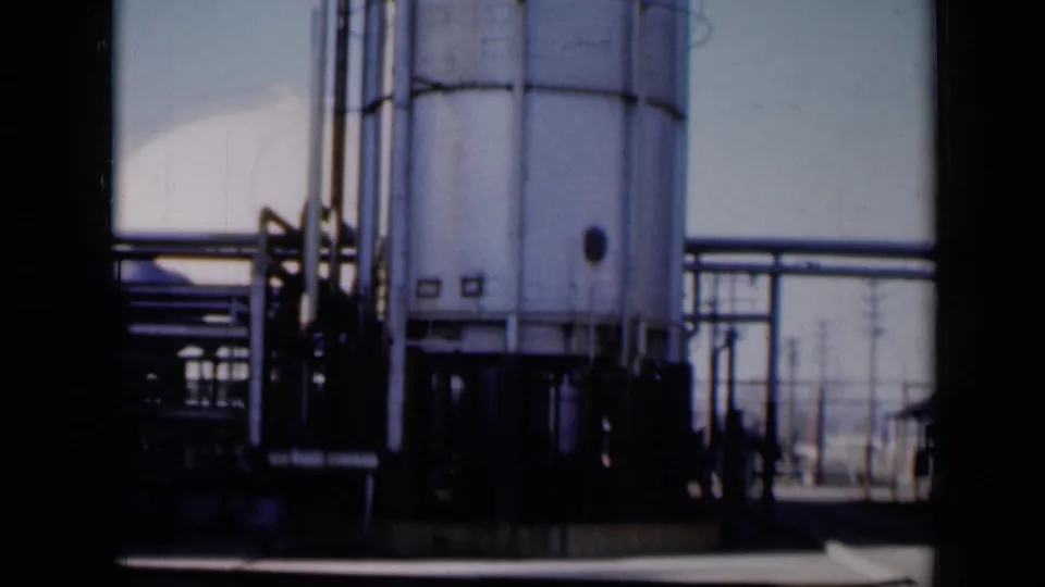 The camera shifts lower on the Missouri industrial site, showing the tank base, pipe runs, railings, and open paved area.