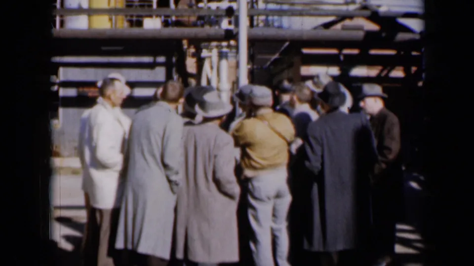 Men in 1960s coats, hats, and workwear stand near Missouri factory equipment, appearing to inspect the exterior structure.