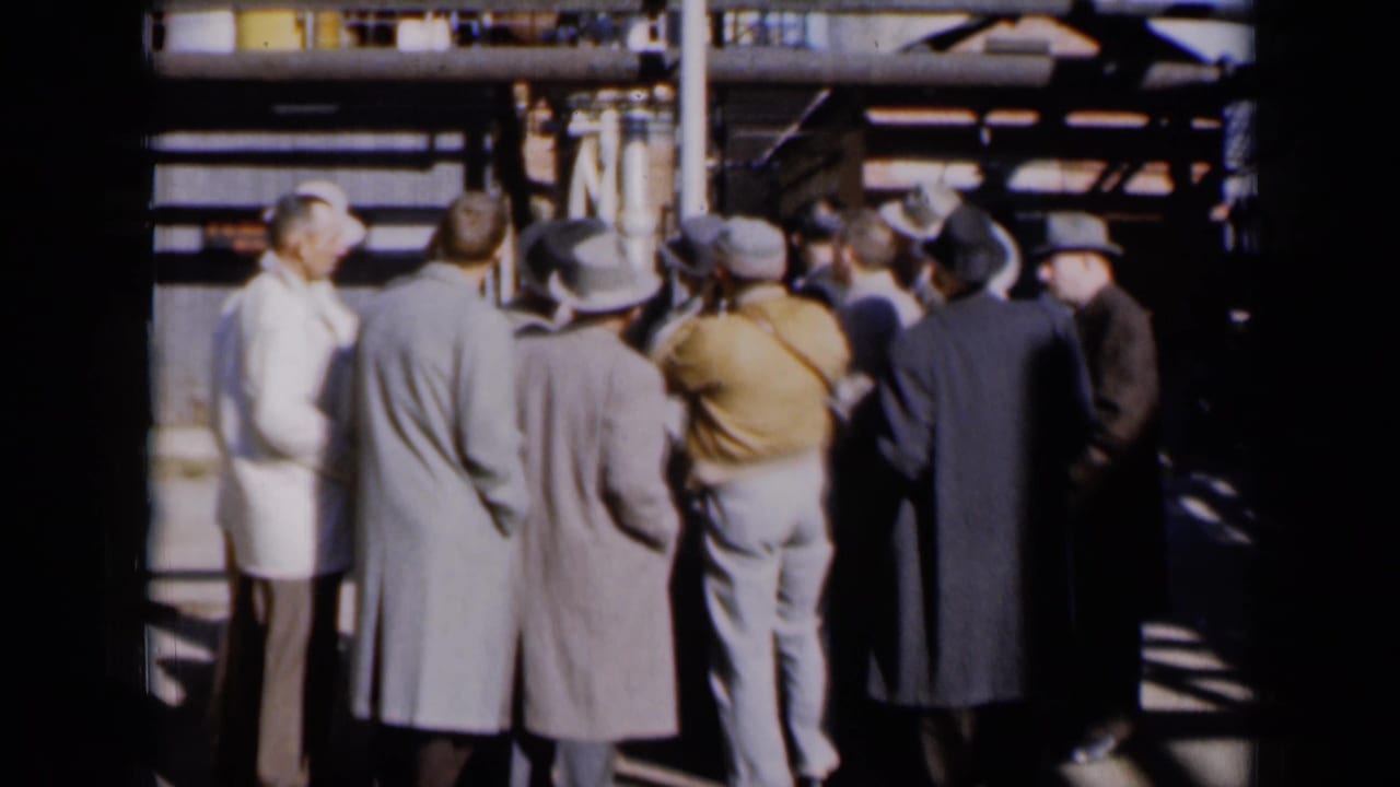 Men in coats and hats gather near industrial pipework while a man in a white coat faces them.