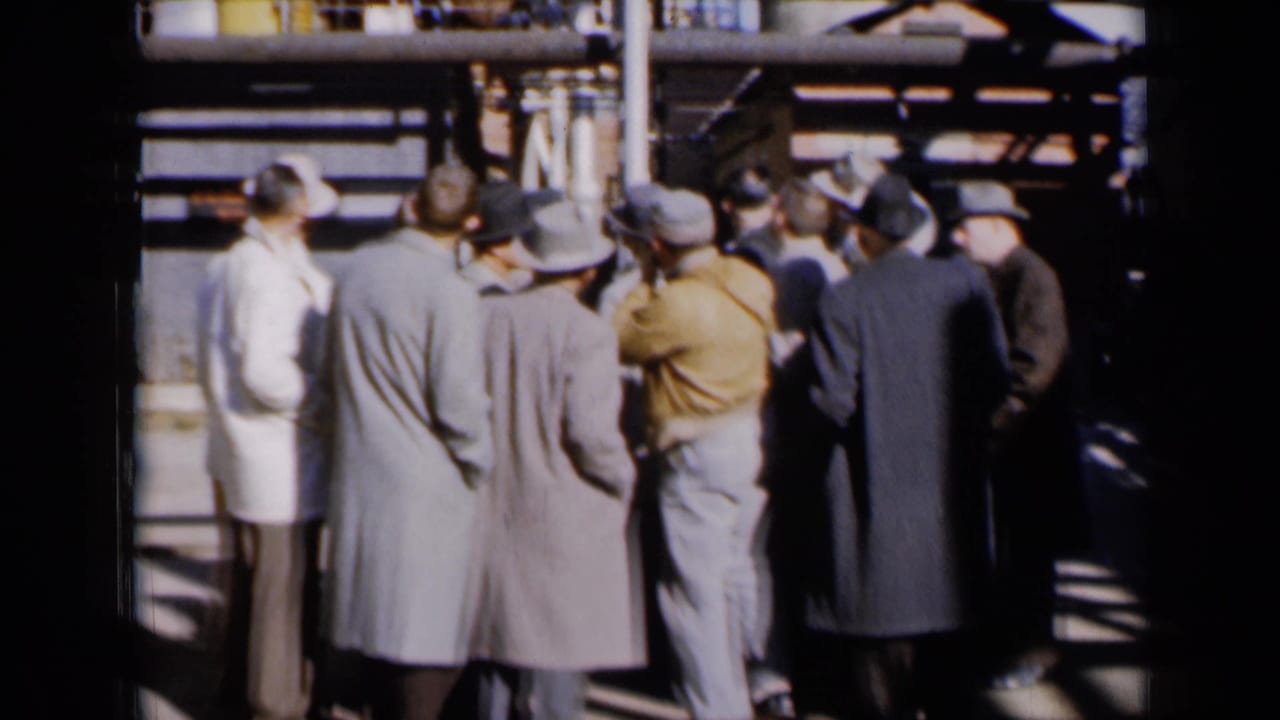 A small group of men stands in a circle near pipes, with one figure gesturing upward.