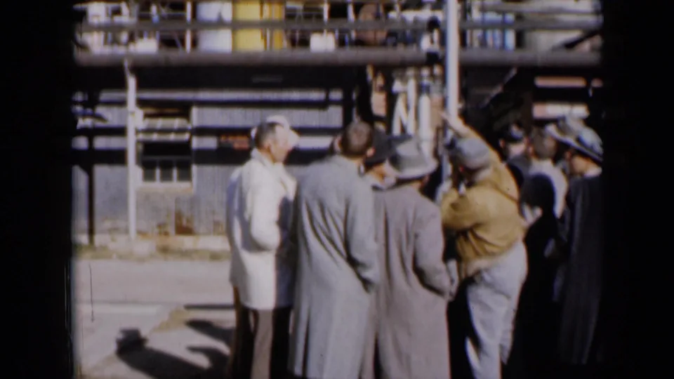 1961 Missouri 8mm frame of men viewed from behind during an outdoor industrial facility discussion in bright sunlight.