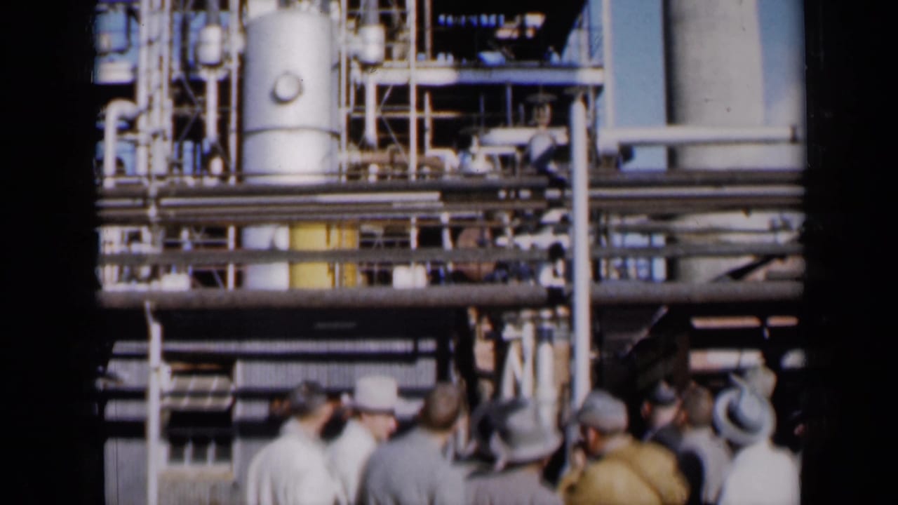 Men stand near a large industrial plant with pipes, tanks, and scaffolding.