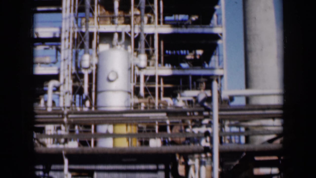 Industrial plant machinery with pipes, tanks, platforms, and vertical stacks.
