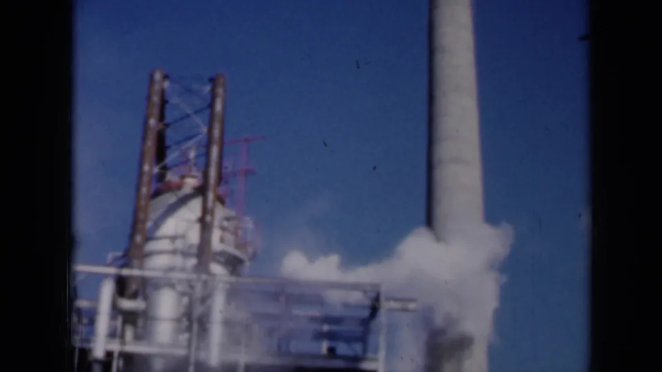 1961 Missouri industrial frame with a vertical smokestack, scaffolded equipment, and a low white plume crossing the foreground.