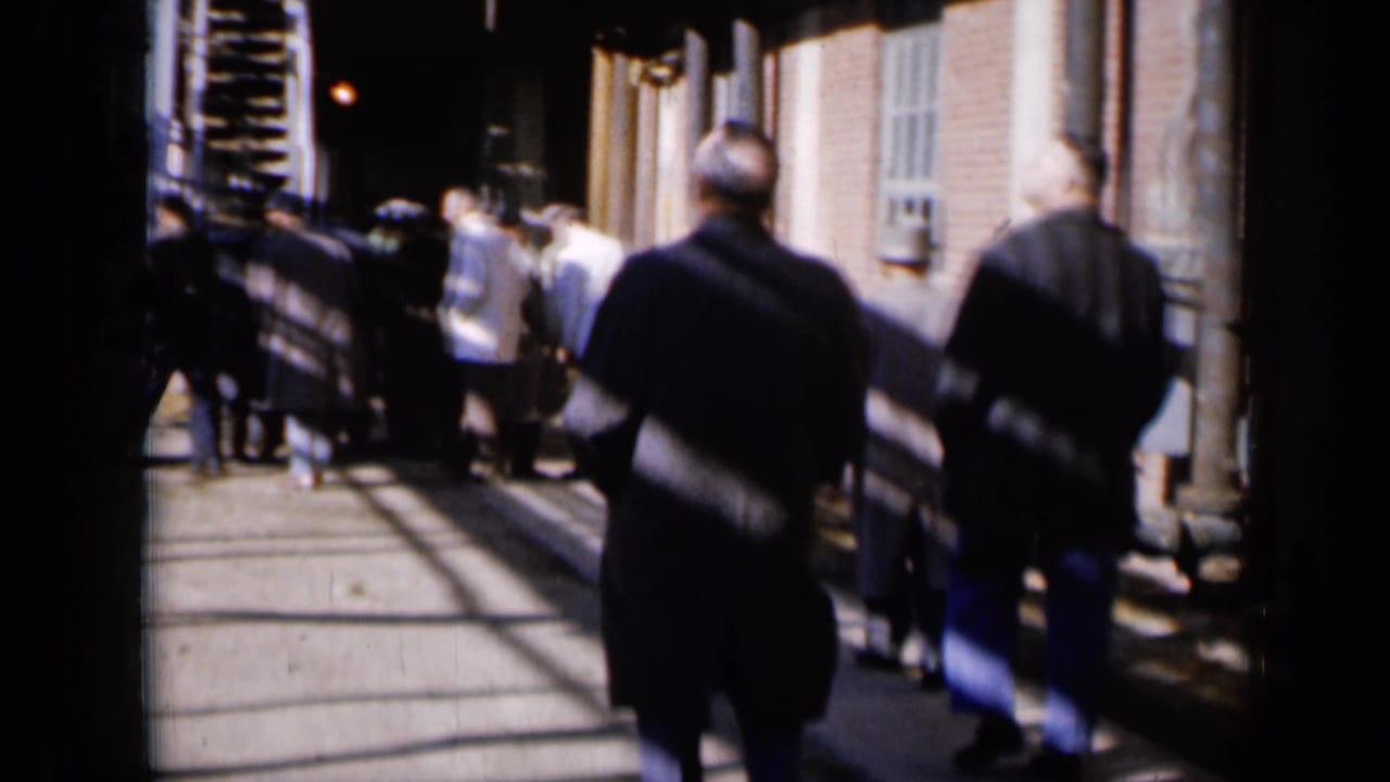 Men walking away through a sunlit alley beside brick buildings and exterior stairs.