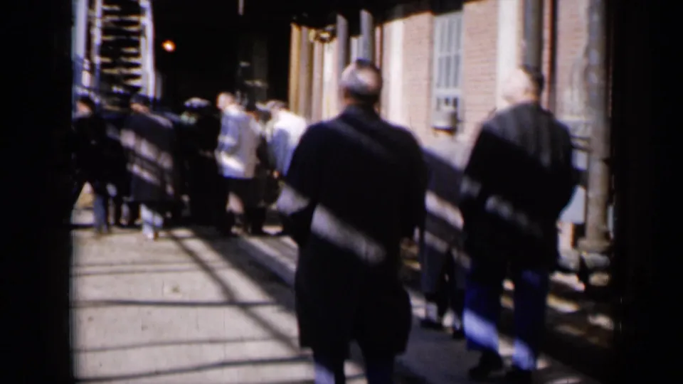 Men in dark coats walk through a Missouri industrial alley in 1961, with brick walls, stairs, and strong shadows visible.