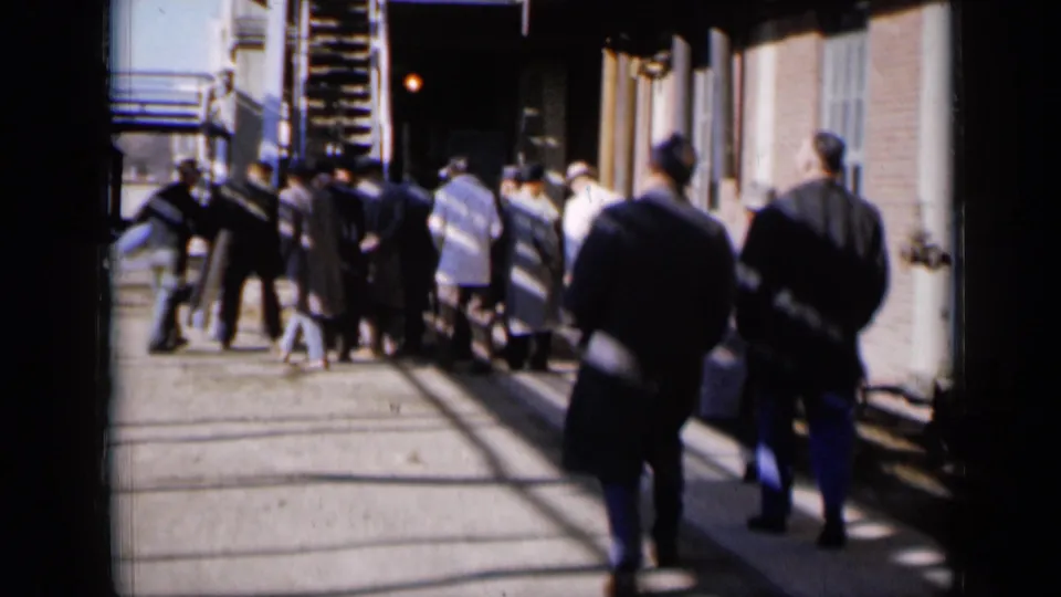 A handheld 1961 Missouri frame shows men walking away past brick industrial buildings and exterior metal stairs.