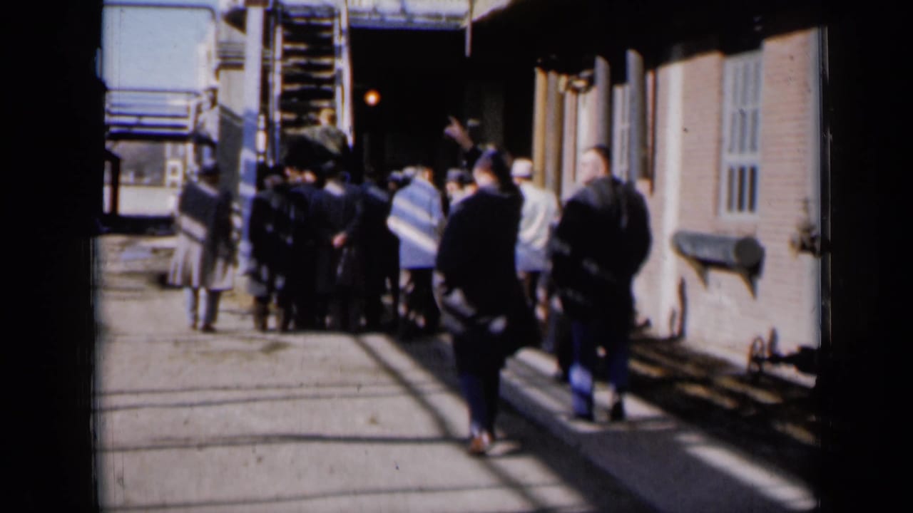 In Missouri 1961 home-movie footage, men in dark coats and hats cluster near an exterior stairway beside a brick building.