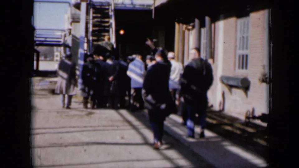 In Missouri 1961 home-movie footage, men in dark coats and hats cluster near an exterior stairway beside a brick building.