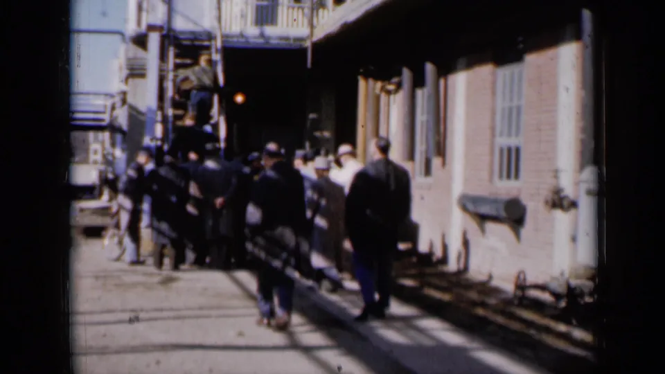 Men in period coats stand and shift toward an exterior metal stairway in a Missouri 1961 home-movie scene.