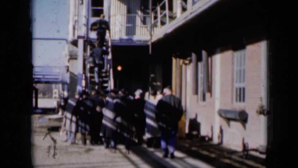 Men remain gathered below an exterior metal staircase in Missouri 1961 footage, with one person visible on the stairs above.