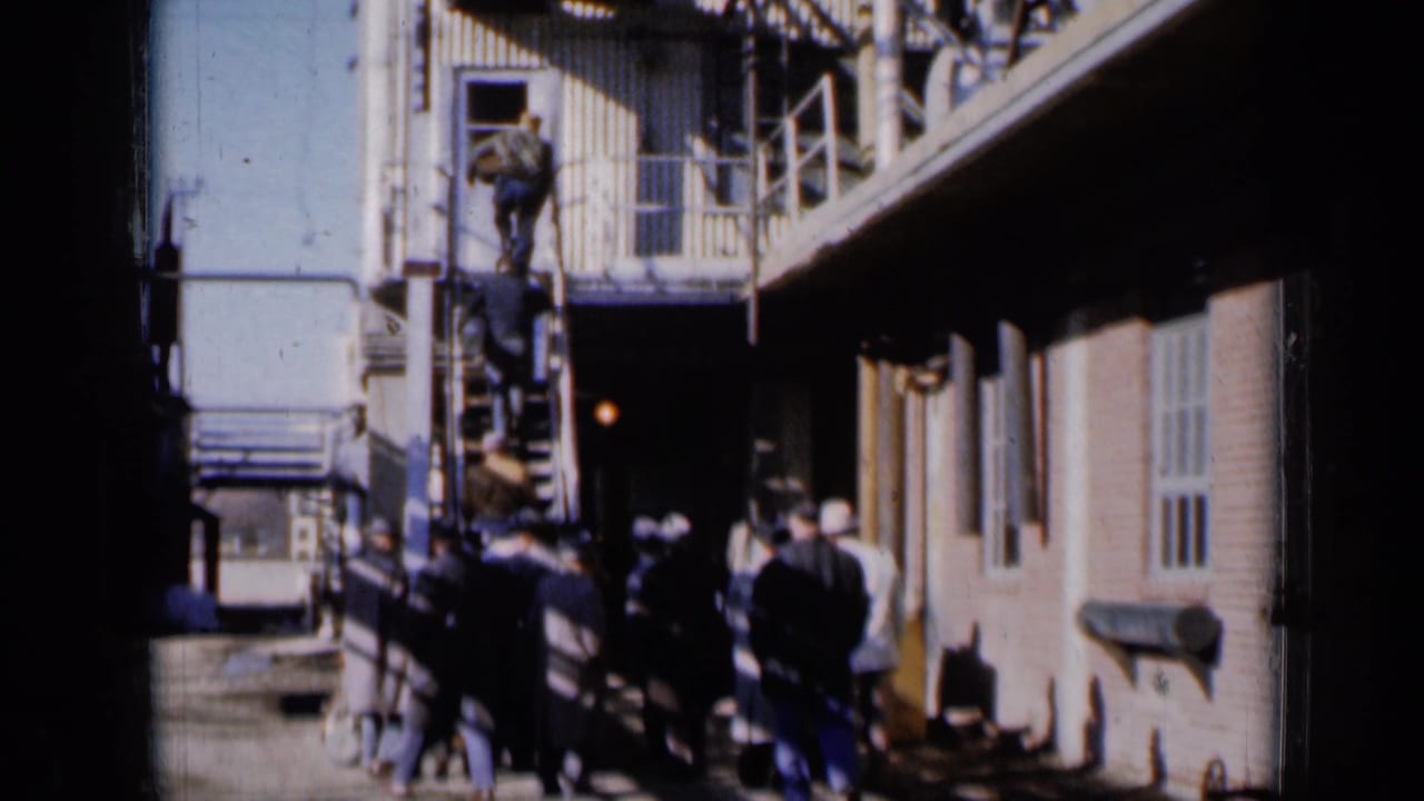 1961 Missouri frame of adults gathered near an exterior metal stair beside a brick community building.