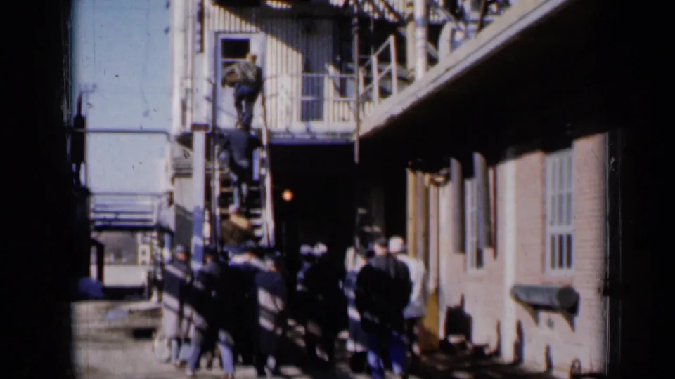 1961 Missouri frame of adults gathered near an exterior metal stair beside a brick community building.