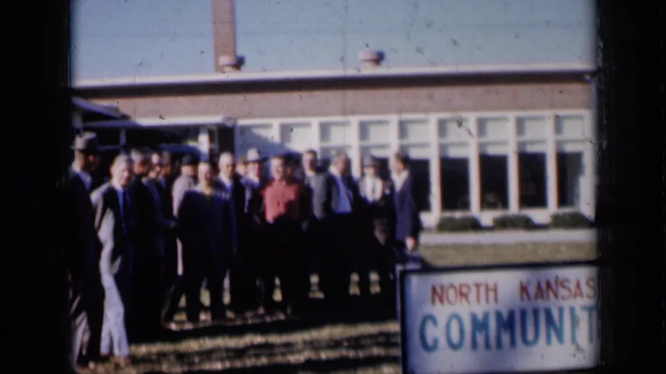1961 Missouri home-movie frame with adults outside a community building and a partly visible North Kansas Community sign.