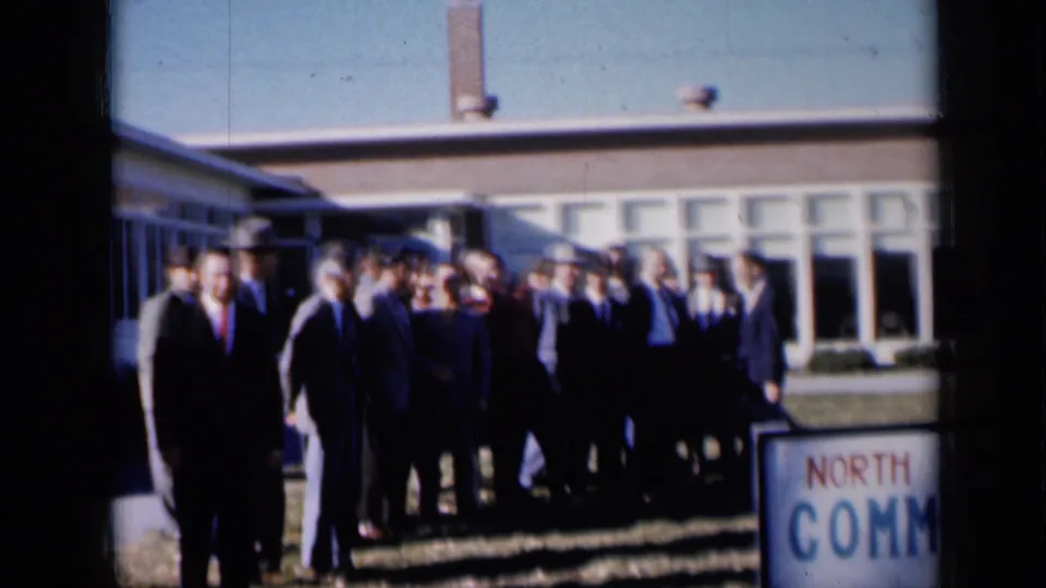 1961 Missouri frame showing a suited group outside a community building with a cropped North Kansas Community sign at right.
