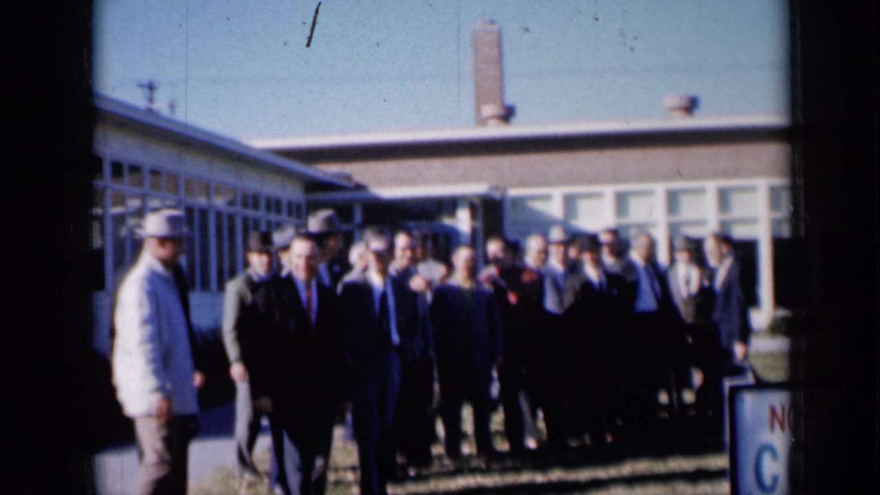 Men in suits and hats gather outside a low building with large windows.